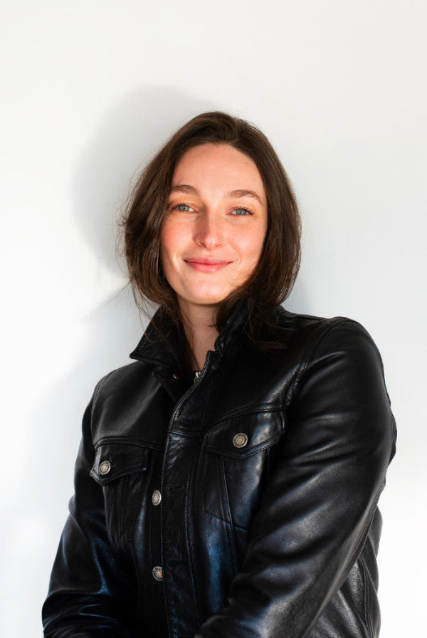 Portrait of a Ilana, founder of Cook For Your Crush  wearing a black leather jacket smiling against a white wall, shot by photographer Claudio Fleitas